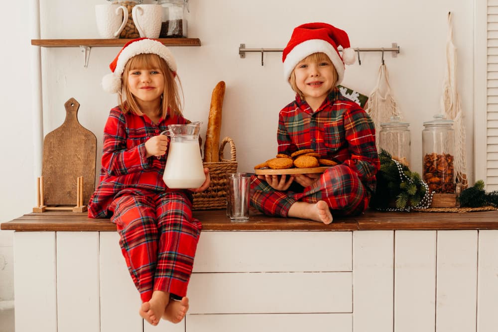 Two kids sit on a kitchen counter. They’re wearing matching red - plaid Christmas pajamas and Santa hats. 